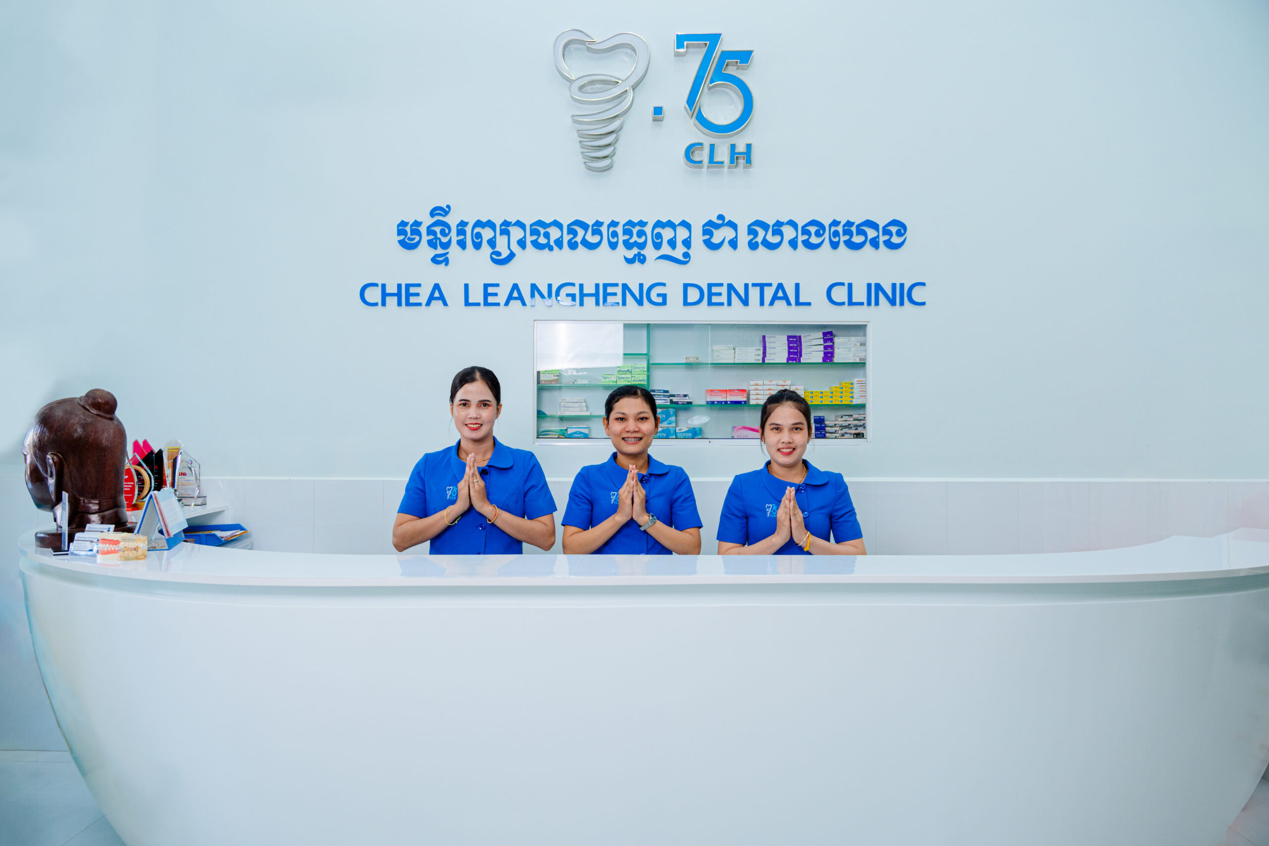 Front desk staff at Chea Leangheng Dental Clinic in blue uniforms, smiling and greeting with traditional Cambodian gesture in front of the clinic logo.