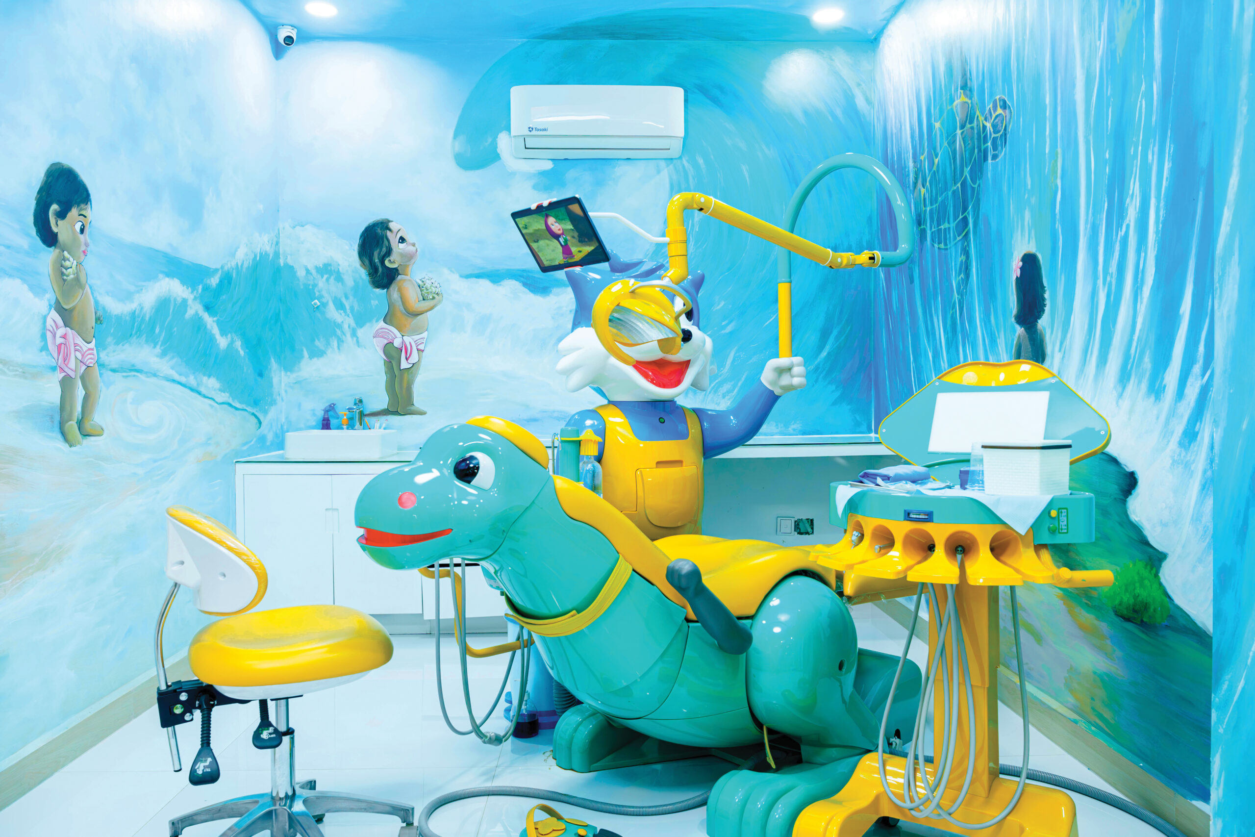 A pediatric dental treatment room with a blue ocean wave mural, featuring a child-friendly dinosaur-shaped dental chair in teal and yellow, and a cartoon cat character holding a tablet.