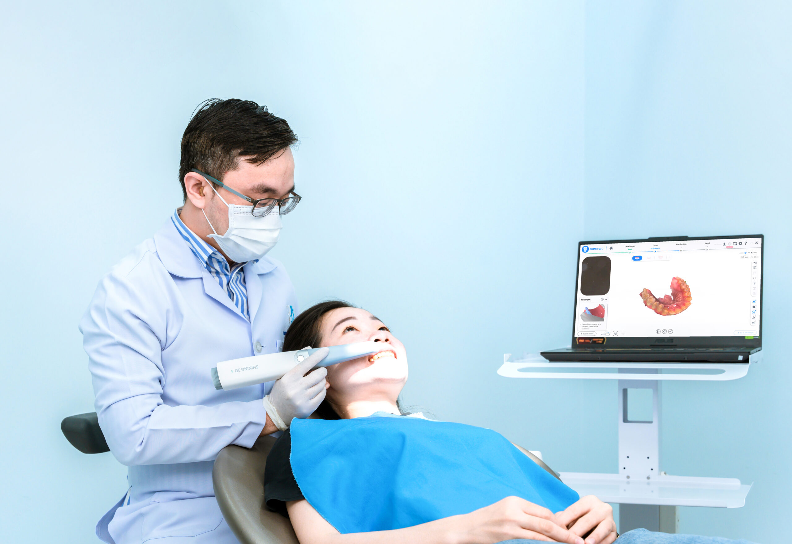 A dentist wearing a face mask and gloves using a handheld digital intraoral scanner to create a 3D dental model of a patient's teeth, with the real-time digital scan displayed on a laptop screen nearby.