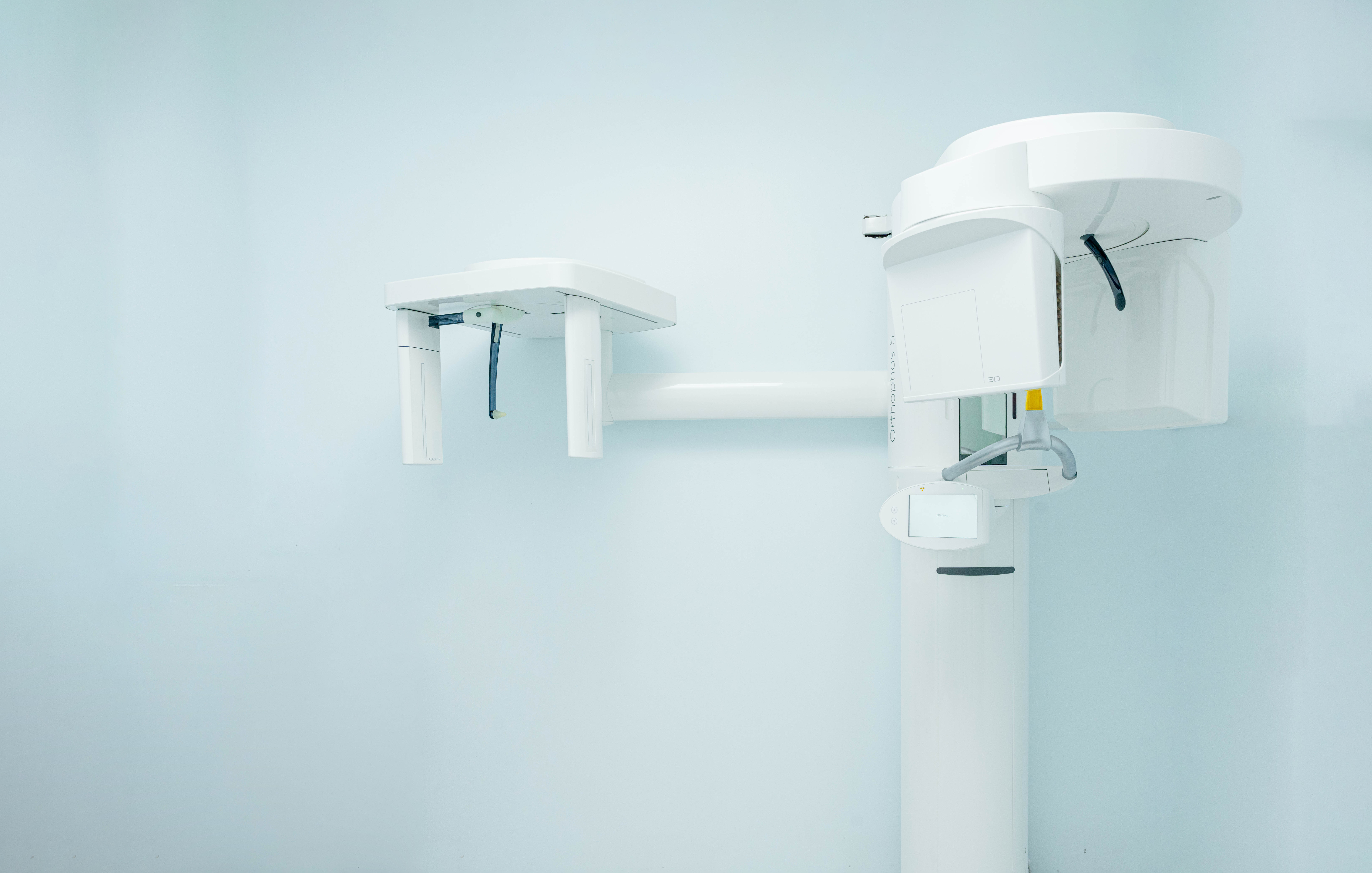 A modern white 3D panoramic dental X-ray and cephalometric machine mounted against a clean, light blue wall in a dental clinic.